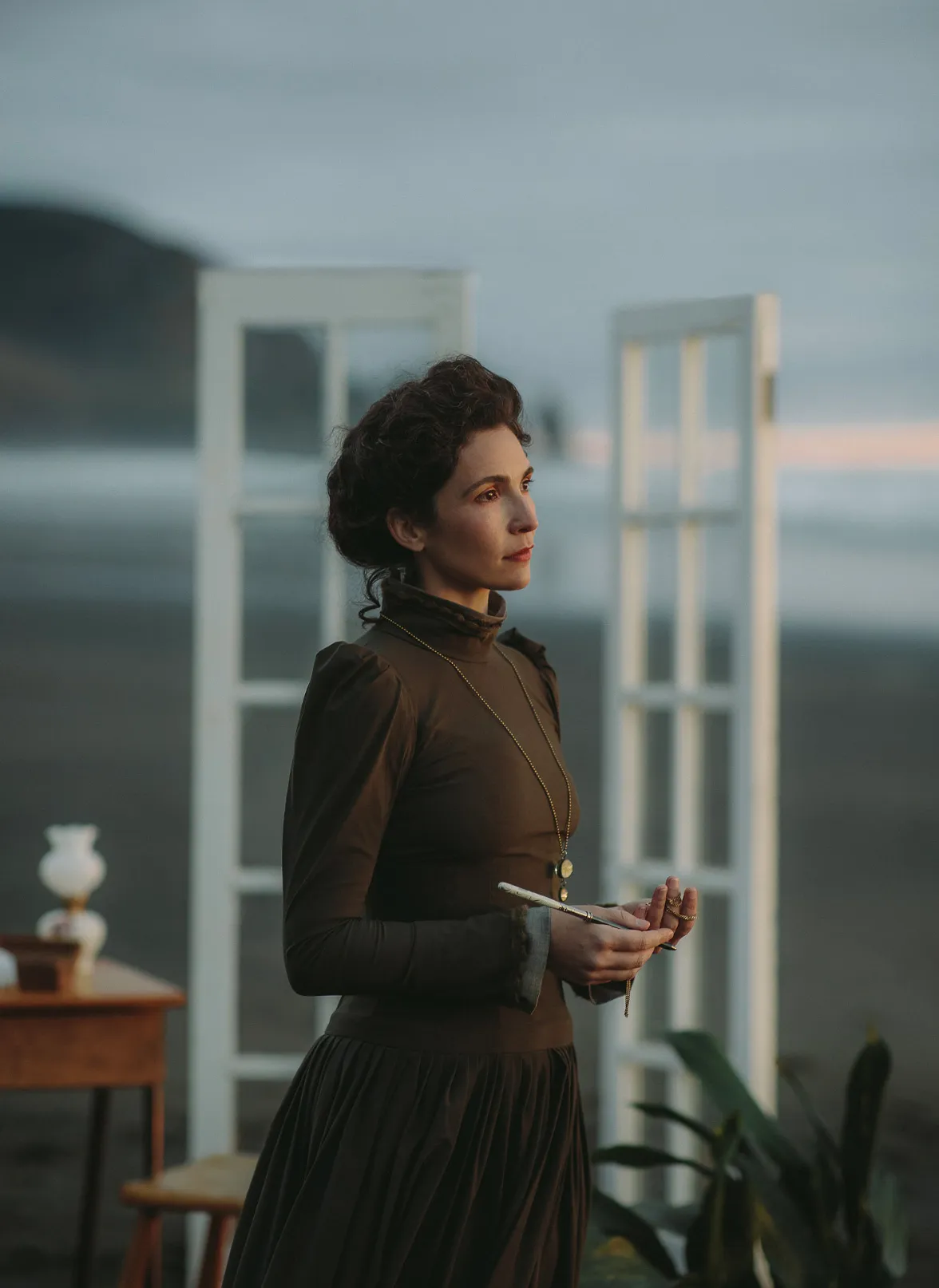 A woman in a dark high-necked period costume standing by a window