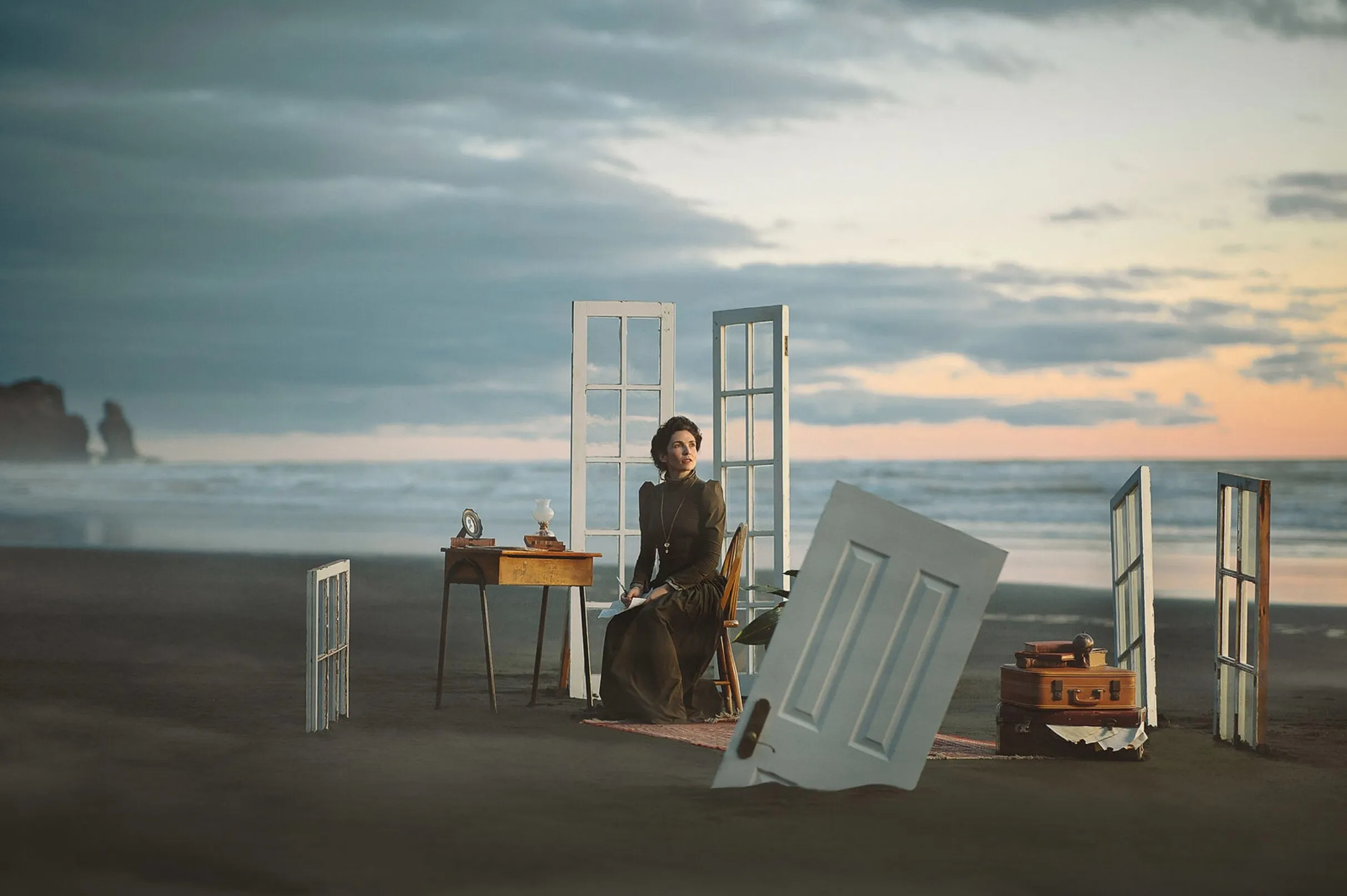 Wide view of the character seated at a writing desk on a beach, surrounded by standing window frames and a fallen door