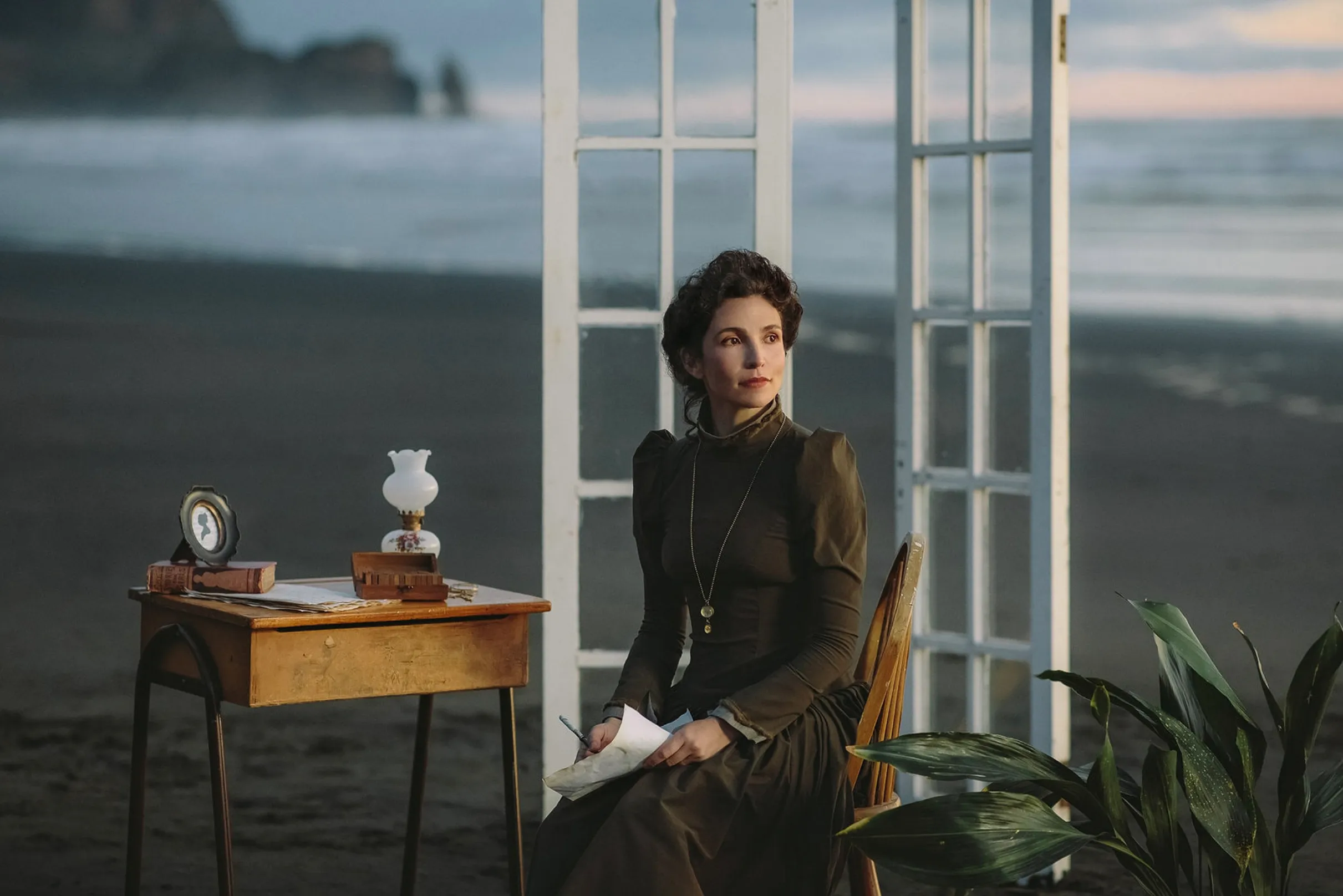 The character seated at a desk on the beach, holding a letter, framed by white windows