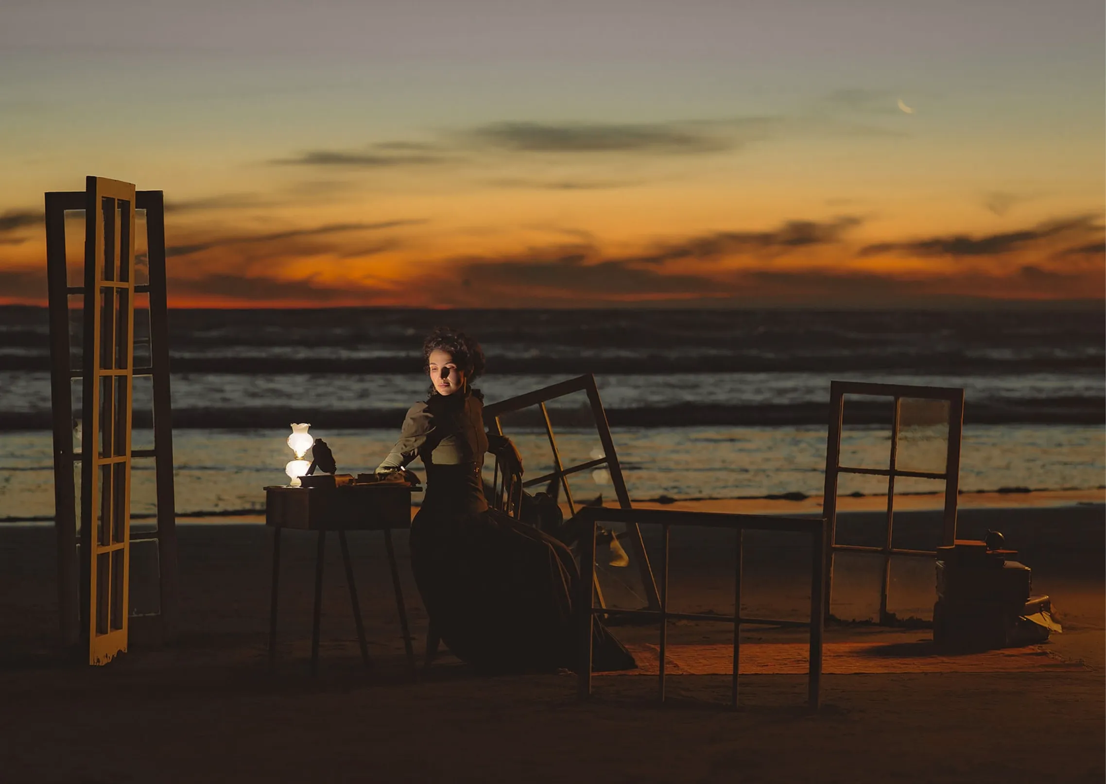 A woman in a dark period costume seated at a writing desk on Piha beach at sunset, surrounded by standing window frames
